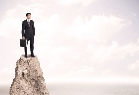 Successful sales person with brief case standing on top of a mountain cliff edge looking above the landscape between the clouds