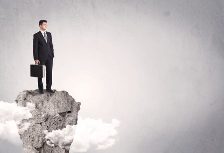 A happy successful businessman standing on a stone cliff with clouds in front of clear empty grey background concept