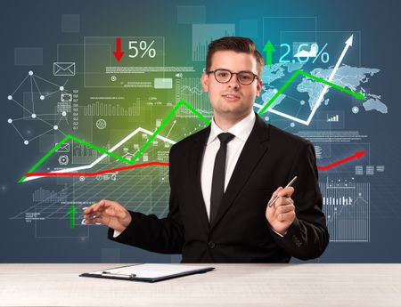 Young handsome businessman sitting at a desk with stocks and progress charts behind him