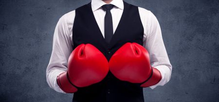 A well dressed sales person standing with red boxing gloves on his hand in front of urban grey wall background concept.