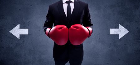 A confident businessman standing with red boxing gloves on his hand in front of arrows pointing in different directions on urban wall background concept.