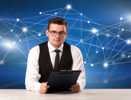 Young handsome businessman sitting at a desk with a blue connection graphic behnid him 