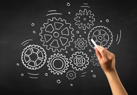 Female hand holding white chalk in front of a blackboard with gears drawn on it