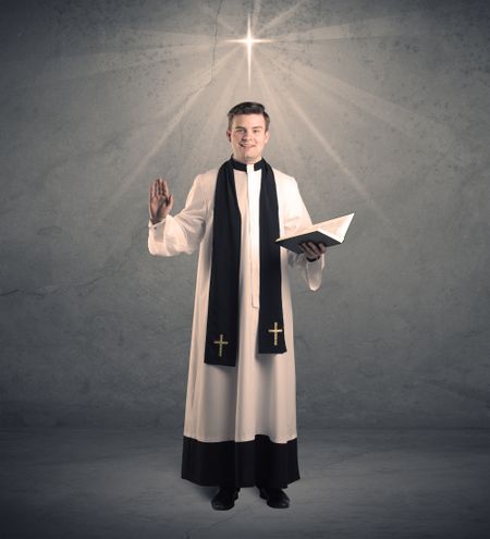 A young male priest in black and white giving his blessing in front of grey wall with glowing cross concept.