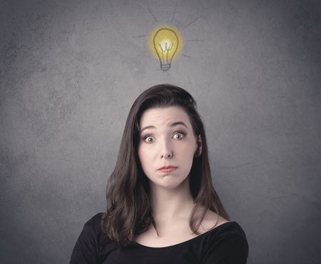 A teen student girl with funny facial expression has a good idea illustrated by a drawn light bulb lighting up above the head on the grey wall background concept.