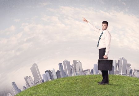 A successful male good looking business man standing in small green grass in front of city landscape with skyscrapers concept.
