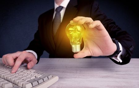 A business man in suit holding a glowing yellow light bulb in his hand while working in the office concept.