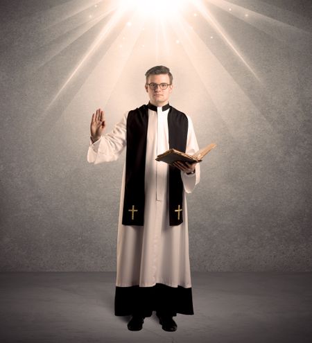 A male religious young priest in black and white dress giving his blessing, holding the holy bible while being illuminated from strong light beams coming from above concept