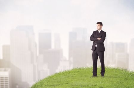 A cheerful caucasian elegant sales manager standing in small green grass in front of faded city landscape, tall buildings concept.