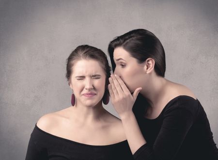 Two fancy dressed actress girls with long hair and make up whispering in front of grey urban background concept.