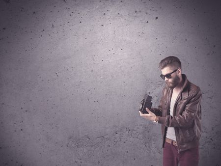 A stylish funny hipster person holding a vintage camera and taking photographs in front of a concrete clear empty urban wall background concept