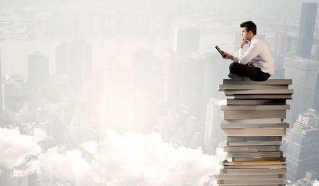 A serious businessman with laptop tablet in elegant suit sitting on a stack of books in front of cityscape