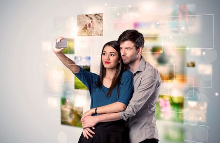 Collecting memories concept with photos in backbround and a happy young couple in love taking selfie with a mobile phone