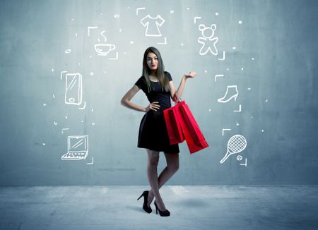 A beautiful young girl in black standing with red shopping bags in front of urban wall background and laptop, shoes, tennis icons concept