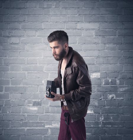 A stylish hipster guy with beard and sunglasses standing in casual clothes in front of an urban blue brick wall background concept