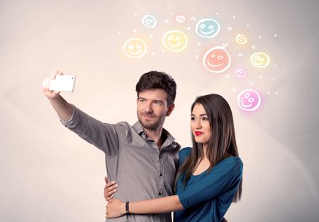 A cheerful young couple taking selfie photo with mobile phone and colorful happy smiley faces illustration above them concept