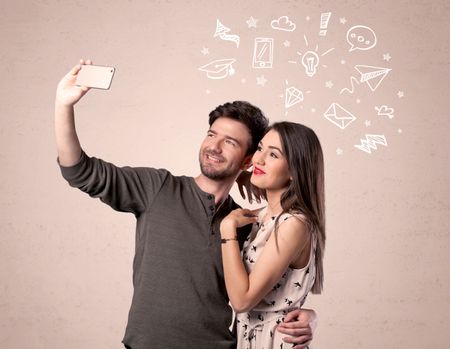 A young couple in love taking selfie with a mobile phone in the handsome guy's hand and drawn media communication icons above them, confused ideas concept