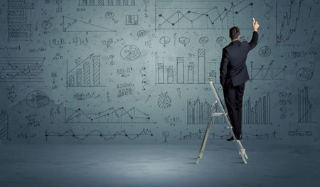 A businessman in modern stylish elegant suit standing on a small ladder and drawing pie and block charts on grey wall background with exponential progressing curves, lines, circles, angles,  numbers