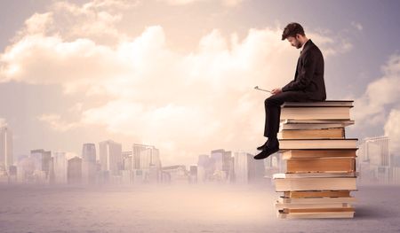 A serious student with laptop tablet in elegant suit sitting on a stack of books in front of cityscape with clouds