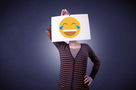 Young casual woman hiding behind a laughing emoticon on cardboard
