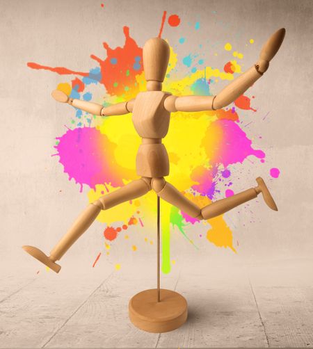 Wooden mannequin posed in front of a greyish background with colorful splashes behind it