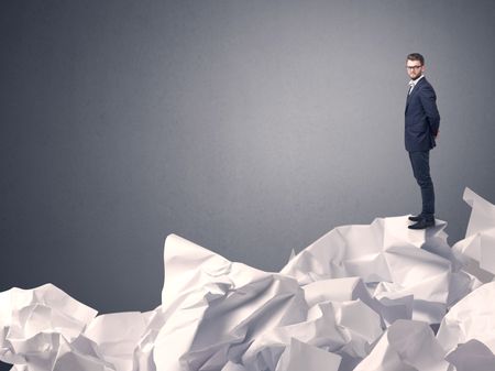 Thoughtful young businessman standing on a pile of crumpled paper with a grey background