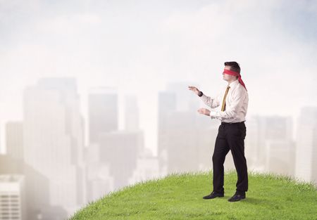 Young blindfolded businessman steps on a a patch of grass with a city in the background