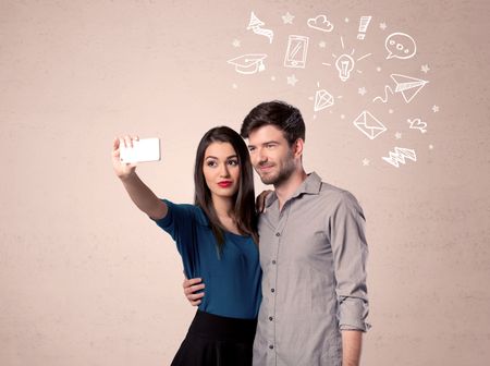 A young couple in love taking selfie with a mobile phone in the handsome guy's hand and drawn media communication icons above them, confused ideas concept