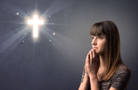 Young woman praying on a grey background with a shiny cross above her