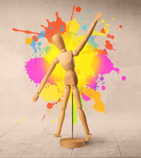 Wooden mannequin posed in front of a greyish background with colorful splashes behind it