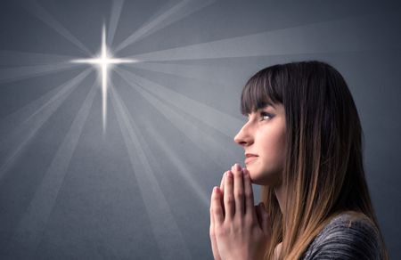 Young woman praying on a grey background with a shiny cross silhouette above her