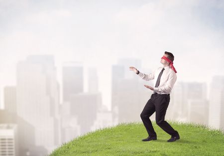 Young blindfolded businessman steps on a a patch of grass with a city in the background