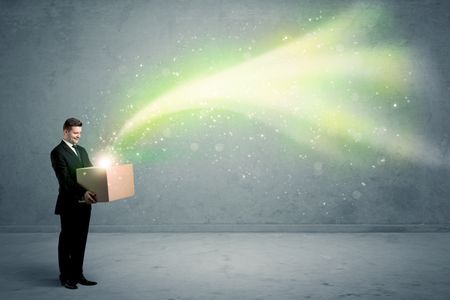 Bright yellow, green light beams escaping a cardboard box held by young elegant male business person in stylish suit concept.