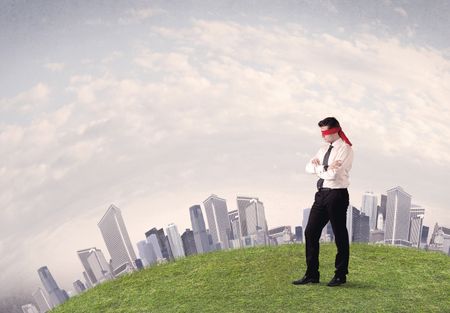 Young blindfolded businessman steps on a a patch of grass with a grey buildings in the background