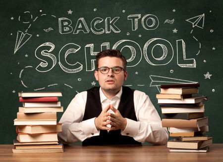 A young teacher in glasses sitting at classroom desk with pile of books in front of blackboard saying back to school drawing concept.