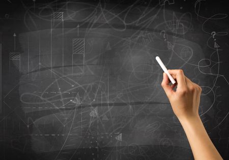 Female hand holding white chalk in front of a blackboard with scribbles and plans drawn on it 