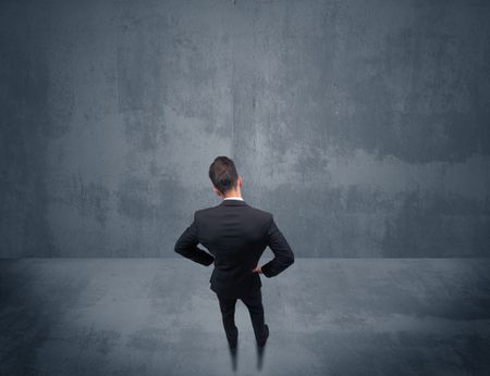 A young businessman with briefcase standing in blank empty space facing a grey urban wall, scratching his head concept.