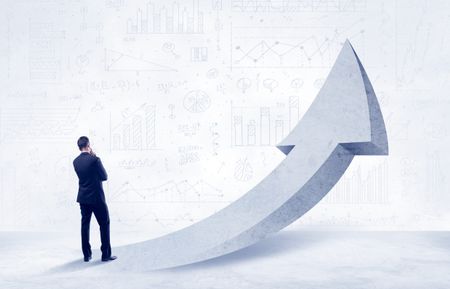 Young sales business person in elegant suit standing with his back in front of a big arrow pointing up and a clear background full of pie charts, numbers