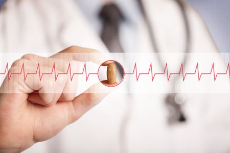A male doctor in white coat with a stethoscope on one shoulder holding a pill between his fingers focused on a heartbeat graph