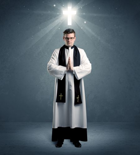 A young caucasian priest with deep faith blessing with the holy bible in his hand in front of blue wall background with illustrated glowing cross concept.