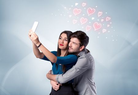 young-couple-love-drawn-red-hearts-675499435 A young couple in love and drawn red hearts taking selfie with a mobile phone in the handsome guy's hand in front of an empty clear grey wall background concept