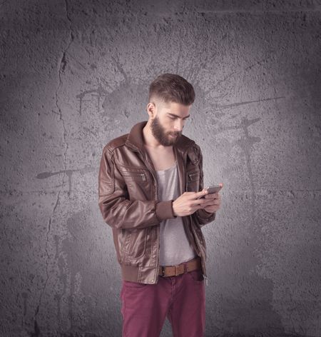 A hipster fashion model guy in casual clothing stnading with mobile phone in front of concrete urban wall background concept