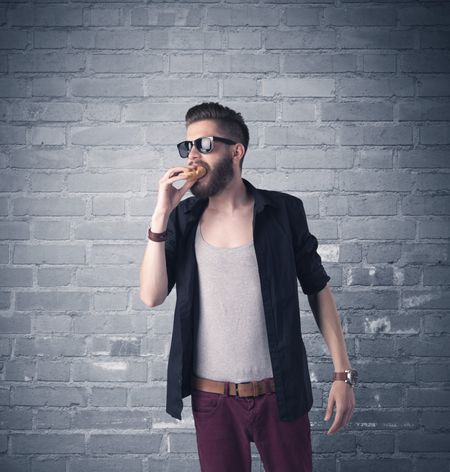 A stylish hipster guy with beard and sunglasses standing in casual clothes in front of an urban blue brick wall background concept
