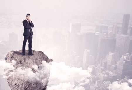 A young successful happy businessman standing confident on stone cliff in the clouds looking at the city scape from above concept
