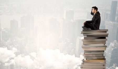 A serious businessman in elegant suit sitting on a pile of giant books in front of a grey city scape with clouds, fog