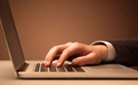 An office worker in elegant suit sitting at desk, typing on portable laptop with empty brown wall background