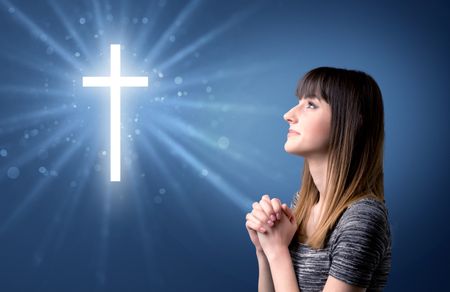 Young woman praying on a blue background with a sparkling cross above her