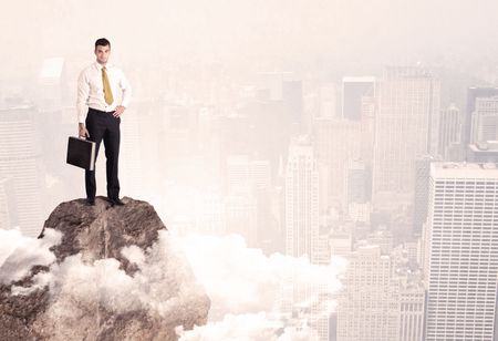 An elegant professional business male standing on top of a high cliff above the clouds looking at the city concept