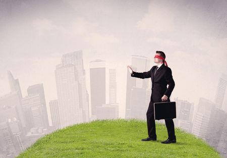 young-blindfolded-businessman-steps-a-patch-678050881 Young blindfolded businessman steps on a a patch of grass with a cloudy city in the background