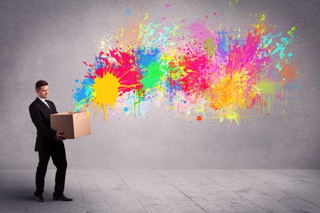 A young smiling business male holding a paperboard box with illustration of colourful spray paint splash on urban wall background concept.